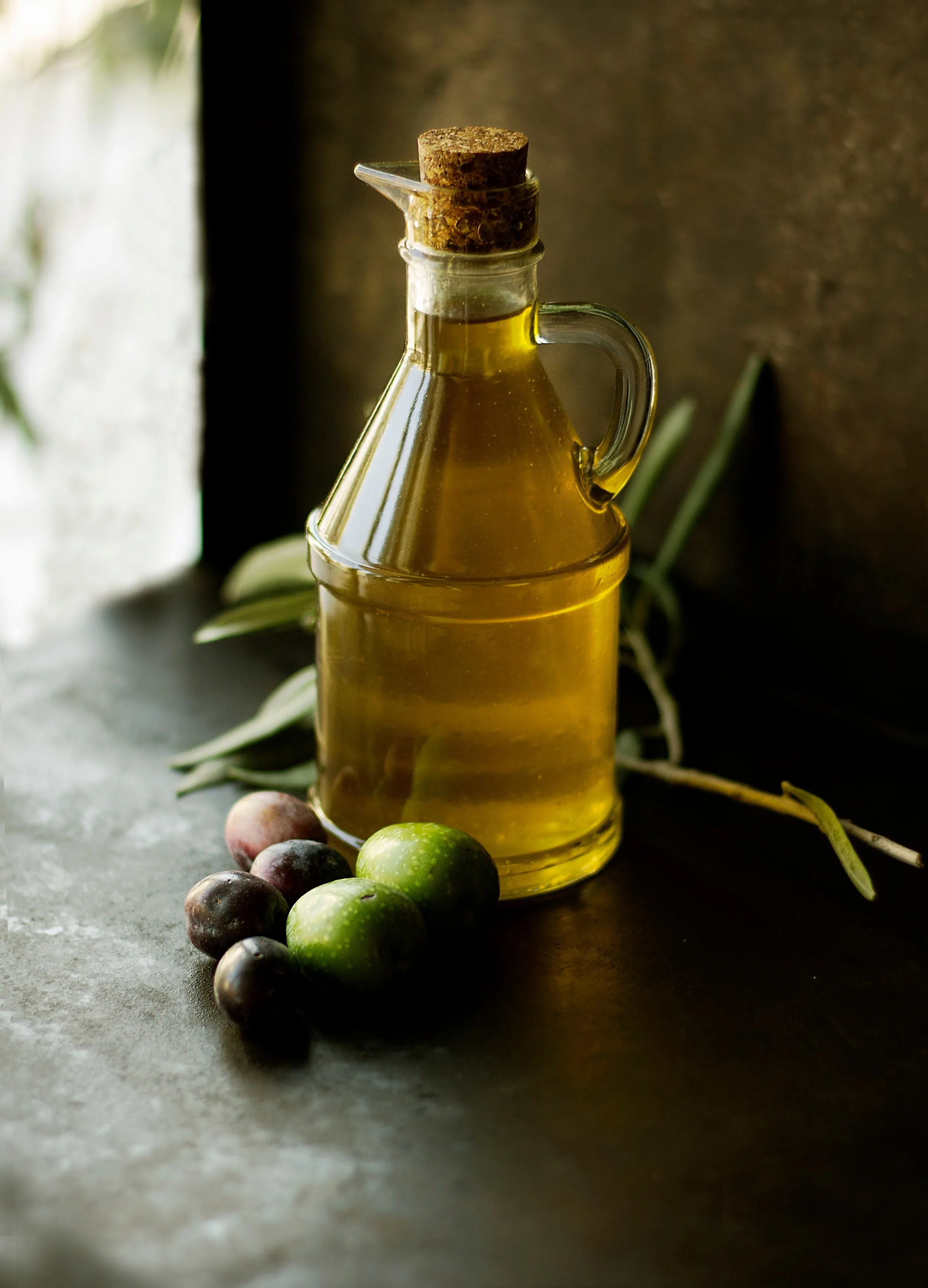 Aceitera de vidrio llena de aceite sobre una encimera, con aceitunas y una rama de olivo al lado